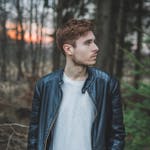 Young man in a black leather jacket standing in a forest during twilight, styled fashion portrait.